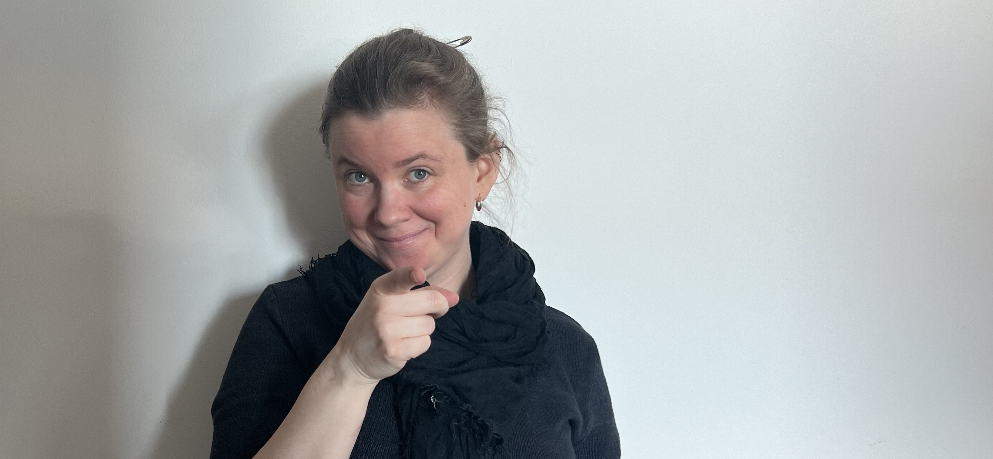 Ester Daniel Ytterbrink on a white background, in black clothes, smiling and pointing into the camera.