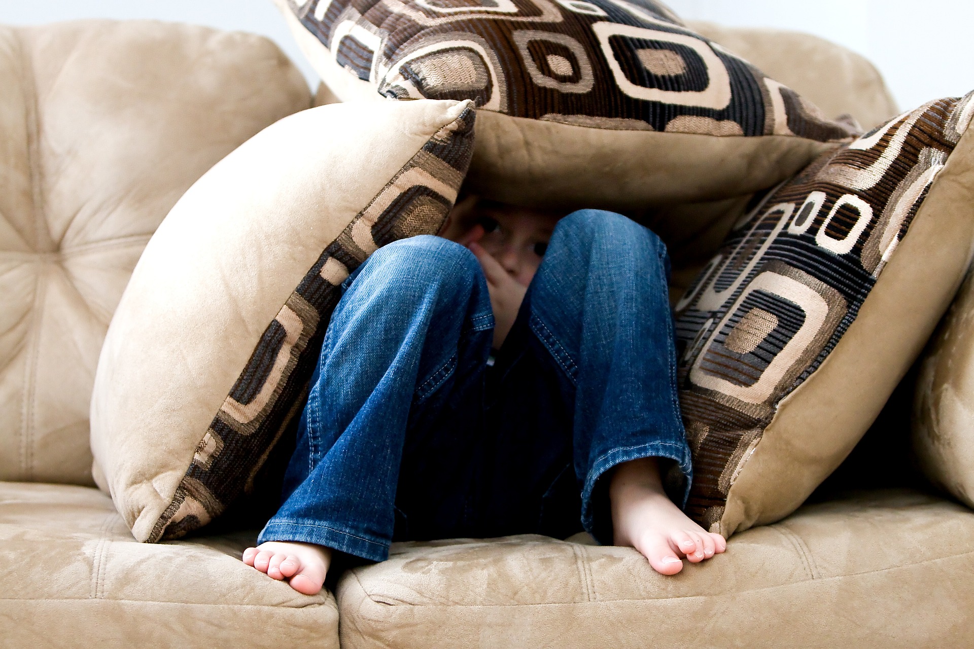 A child sitting in a sofa, surrounded by pillows, legs pulled up, face hardly showing.